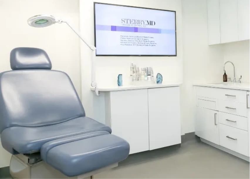 A clean medical exam room with a blue patient chair, overhead exam light, white cabinetry, countertop skincare products, and a wall-mounted screen displaying 
