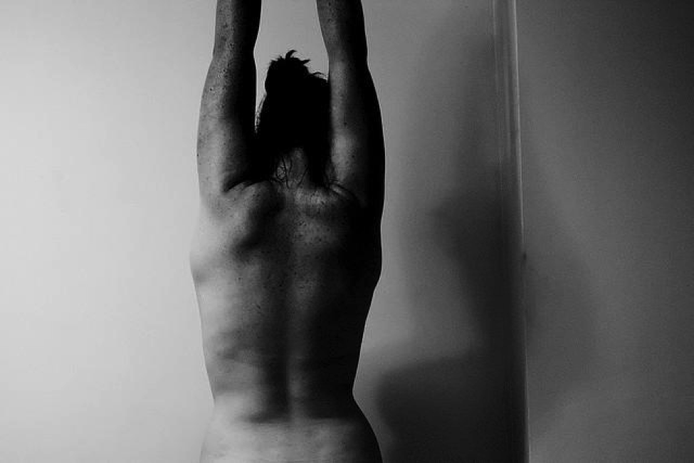 Black-and-white back view of a woman who is looking down with her arms extended straight up over her head.