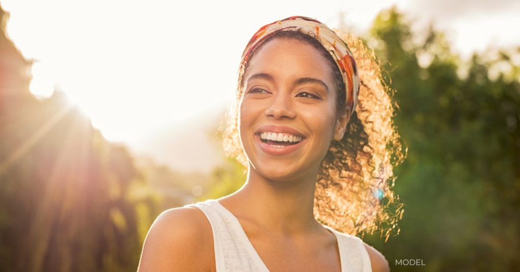 A woman outside in the sunshine smiling with her hair pulled back and wearing a tank top.